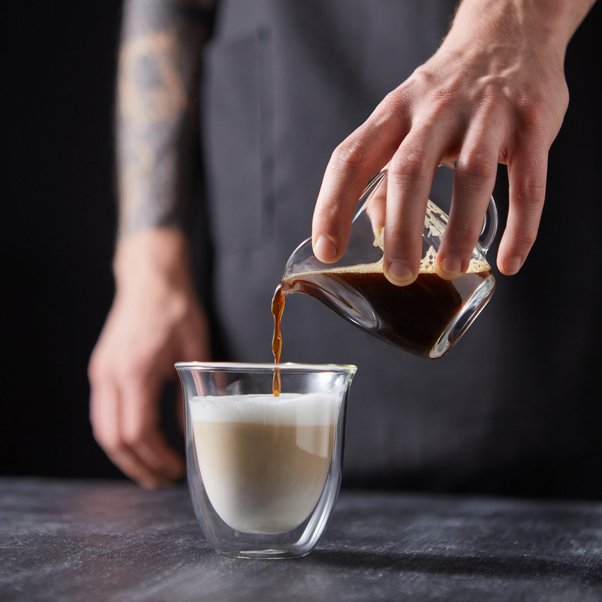 A man’s hand pours coffee into a cup of milk on a black wooden table. Cappuccino preparation.